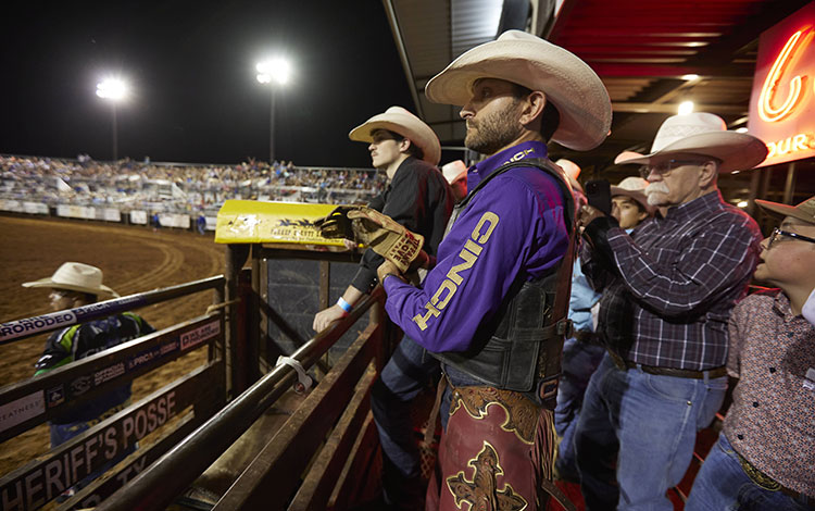 Man wearing a purple shirt and cowboy hat standing on the bucking chutes at a rodeo.